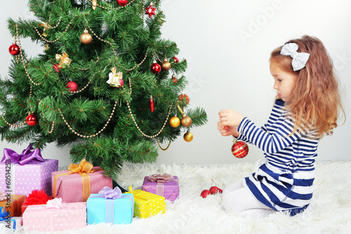 Photo: Little girl decorating Christmas tree with baubles in room Little girl decorating Christmas tree with baubles in room