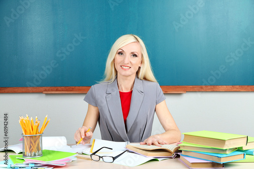 Photo: School teacher sitting at table on blackboard background School teacher sitting at table on blackboard background
