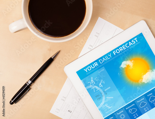 Photo: Tablet pc showing weather forecast on screen with a cup of coffe Tablet pc showing weather forecast on screen with a cup of coffe