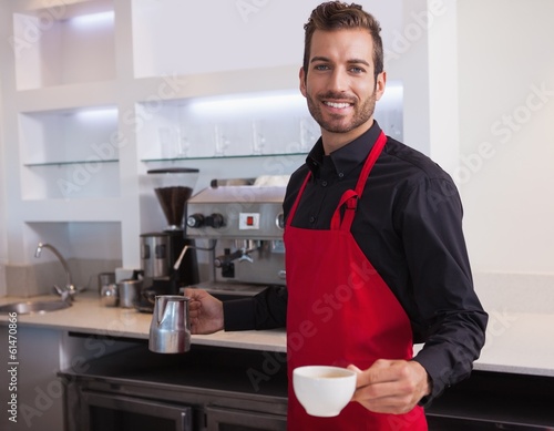 Photo: Handsome young barista holding jug and cup of coffee Handsome young barista holding jug and cup of coffee