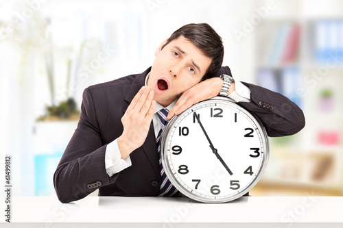 Photo: Sleepy young businessman leaning his head on a big wall clock Sleepy young businessman leaning his head on a big wall clock