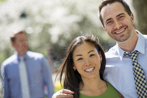 Foto: Three People In Business Dress. A Couple Side By Side Smiling At The Camera. A Man In The Background. Three People In Business Dress. A Couple Side By Side Smiling At The Camera. A Man In The Background.