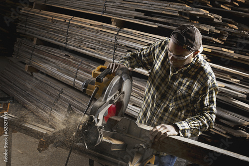 Foto: A reclaimed lumber workshop. A man in protective eye goggles using a circular saw to cut timber. A reclaimed lumber workshop. A man in protective eye goggles using a circular saw to cut timber.