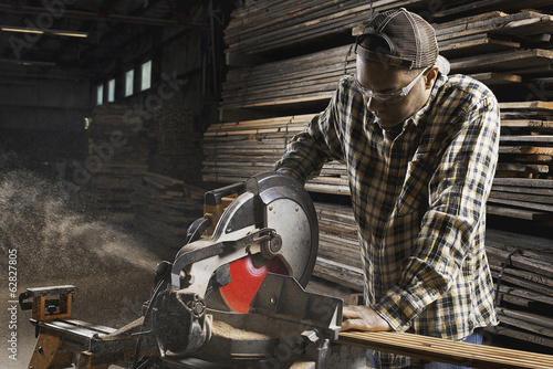 Foto: A reclaimed lumber workshop. A man in protective eye goggles using a circular saw to cut timber. A reclaimed lumber workshop. A man in protective eye goggles using a circular saw to cut timber.