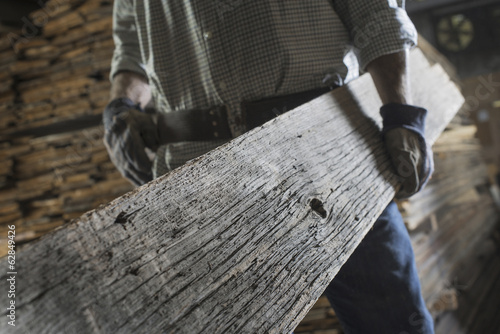 Foto: A heap of recycled reclaimed timber planks of wood. Environmentally responsible reclamation in a timber yard. A man carrying a large plank of mature weathered wood. A heap of recycled reclaimed timber planks of wood. Environmentally responsible reclamation in a timber yard. A man carrying a large plank of mature weathered wood.
