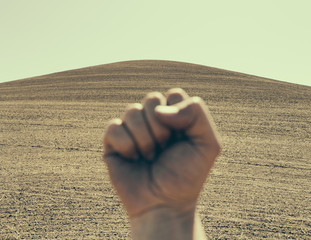 Photo: A hand bunched up and making a fist, in a gesture of defiance or determination, against the background of a ploughed field and farmland. A hand bunched up and making a fist, in a gesture of defiance or determination, against the background of a ploughed field and farmland.