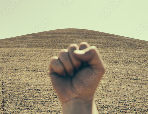 Photo: A hand bunched up and making a fist, in a gesture of defiance or determination, against the background of a ploughed field and farmland. A hand bunched up and making a fist, in a gesture of defiance or determination, against the background of a ploughed field and farmland.