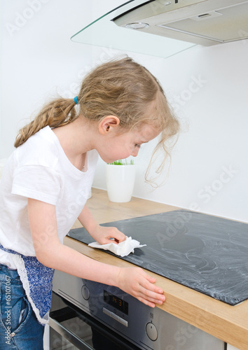 Photo: Little girl wipes cooktop in the kitchen at home. Little girl wipes cooktop in the kitchen at home.