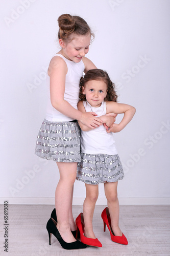 Photo: Beautiful small girls in big shoes on wall background Beautiful small girls in big shoes on wall background