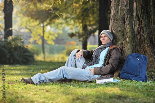 Photo: Male student sitting by a tree in park on a sunny day Male student sitting by a tree in park on a sunny day