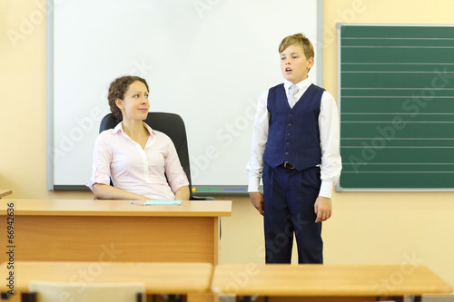 Photo: Teacher sits at table and looks at boy answering near blackboard Teacher sits at table and looks at boy answering near blackboard