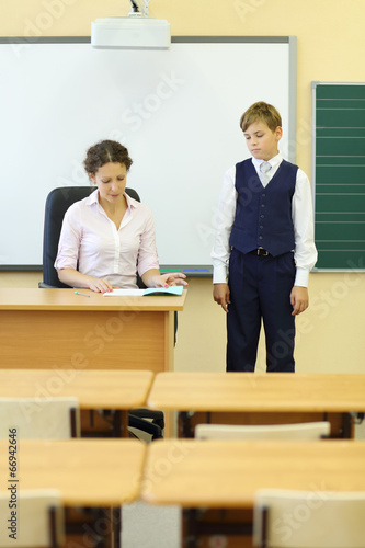 Photo: Teacher sits at table and checks exercise book and boy looks Teacher sits at table and checks exercise book and boy looks