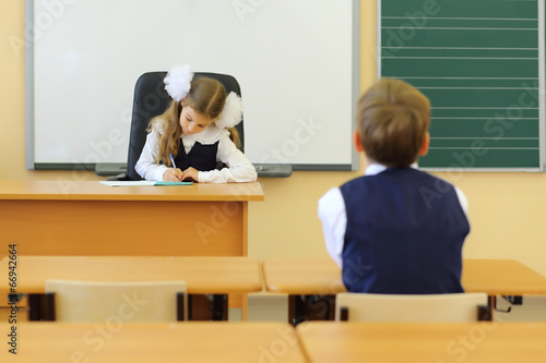 Photo: Girl sits at teacher table and writes and boy sits at desk Girl sits at teacher table and writes and boy sits at desk