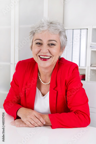 Photo: Portrait: Senior Chefin in Rot sitzend und lachend im Büro Portrait: Senior Chefin in Rot sitzend und lachend im Büro