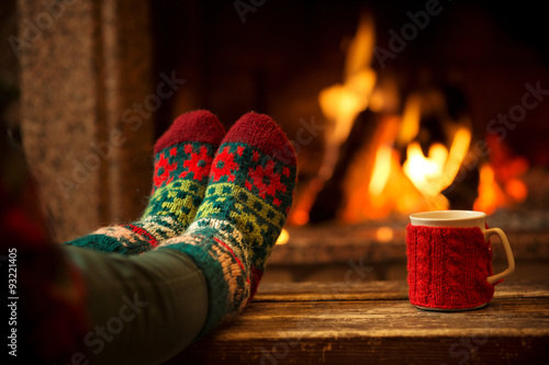 Feet in woollen socks by the Christmas fireplace. Woman relaxes by warm fire with a cup of hot drink and warming up her feet in woollen socks. Close up on feet. Winter and Christmas holidays concept.