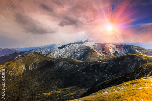 Landscape in the autumn mountains, hills and peaks covered with the first snow. Colorful clouds in the evening sky. Fantastic evening glowing by sunlight.