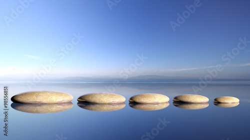  Zen stones row from large to small in water with blue sky and peaceful landscape background.