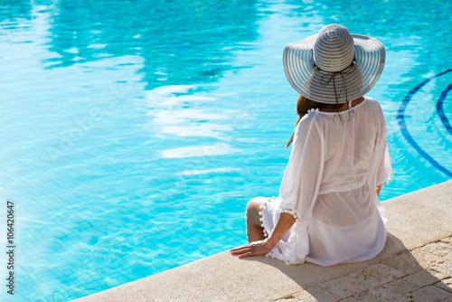 Plakat Tylny widok mody kobieta relaksuje przy luksusowym kurortu zdroju poolside na wakacje. Młoda modna dama jest ubranym słońce kapelusz i białego kaftan.