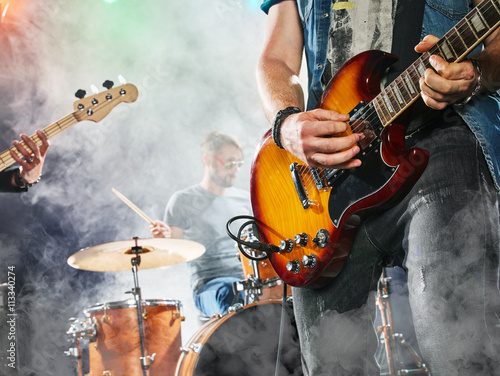 Plakat Zespół rockowy występuje na scenie. Gitarzysta, gitara basowa i perkusja. Gitarzysta na pierwszym planie. Zbliżenie.
