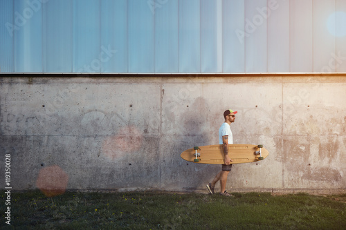 Plakat Hipster w biały t-shirt puste, spodenki, trampki, czapka z daszkiem i okulary przewożących longboard na tle przemysłowych miasta budynku z flary obiektywu