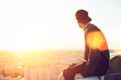 Young and brave man sitting on the edge of the roof and looking far away at the city, lens flares Young and brave man sitting on the edge of the roof and looking far away at the city, lens flares