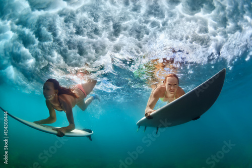 Plakat Grupa aktywnych dziewcząt w akcji. Surfingowiec kobiety z kipieli deską nurkują podwodnego pod łamać dużą fala. Zdrowy tryb życia. Sporty wodne, ekstremalne surfowanie w obozie przygodowym na wakacje rodzinne wakacje plaży