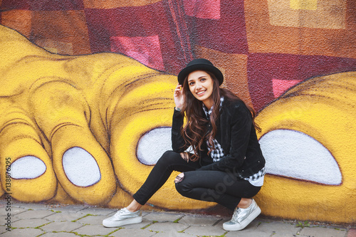 Zdjęcie XXL stylowa młoda kobieta w kapeluszu siedzi w pobliżu ściany z graffiti
