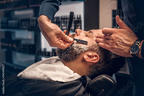 Barber shaving bearded male with a sharp razor. © Fxquadro