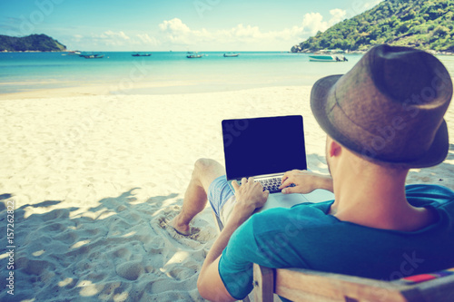 Plakat Atrakcyjny młody człowiek z laptopem pracuje na plaży. Wolność, praca zdalna, freelancer, technologia, internet, podróże i wakacje