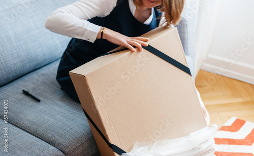 Plakat Młoda piękna kobieta na salonie sofa rozpakowuje unbox pudełko kartonowe zawierające nowe piękne ubrania