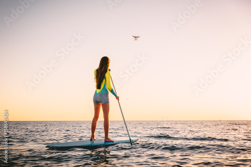 Plakat Atrakcyjna młoda kobieta Stand Up Paddle Surfing i drone copter z pięknymi kolorami słońca