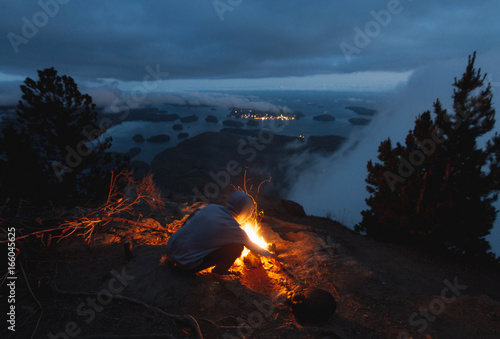 Zdjęcie XXL Man sitting on the edge with campfire above bay with islands and forests in cloudy weather from top view in the evening