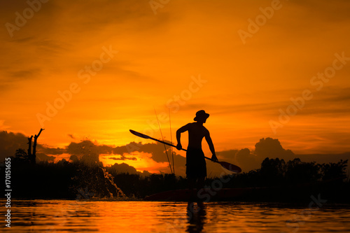 Plakat Sylwetka rybak pozycja na łodzi, chwyta paddle i paddle z pluśnięciem woda na zmierzchu tle.