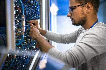 Side view portrait of young man wearing glasses connecting cables in server cabinet while working with supercomputer in data center