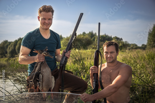 Plakat Rozochoceni mężczyzna myśliwi z pistoletami i trofeum w łodzi na brzeg rzeki podczas łowieckiego sezonu wśród płochy