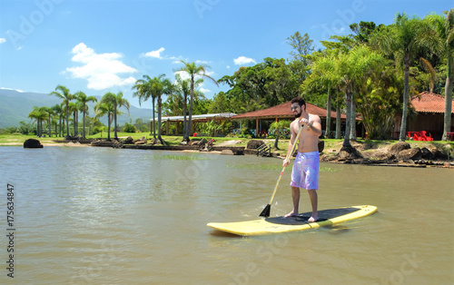 Plakat Młody człowiek uprawiania paddleboarding, wstać wiosła, wiosłowania. Surfing z wiosłowaniem. tło z plaży i palmami.