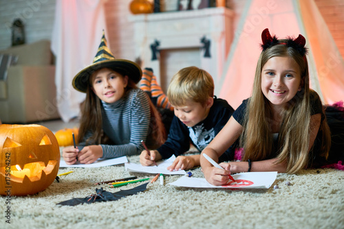Plakat Grupowy portret rozochoceni dzieci jest ubranym Halloweenowych kostiumy zbierał wpólnie przy wygodnym pokojem dekorującym dla Halloween i rysuje kolorowych obrazki, dziewczyny patrzeje kamerę z szerokimi uśmiechami