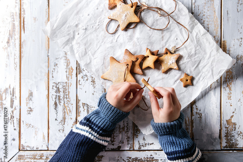 Zdjęcie XXL Dziecko ręki robią girlandzie domowej roboty shortbread gwiazdowego kształta cukrowi ciastka różny rozmiar na nici na pieczeniu tapetują nad białym drewnianym deska stołem. Świąteczny prezent ręcznie robiony. Widok z góry z przestrzenią