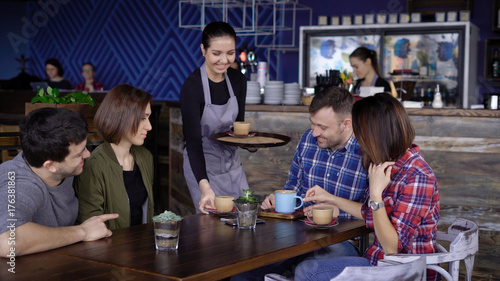 Fotoobraz na płótnie Gościnna młoda kelnerka w kawiarni przynosząc napoje w kolorowych kubkach do klientów. Czterech przyjaciół siedzących w restauracji spędzających wolny czas razem i otrzymując ich zamówienie.