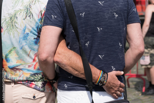 Two gay men stand in the sun at a gay pride festival in Europe. Man puts his arm around his boyfriend's waist. Homosexual couple in love standing outside in town square.