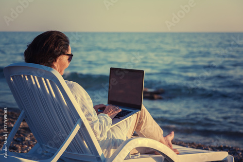 stylish attractive mature woman 50-60 makes use of laptop on the seashore, travel and retirement, fashion and beauty, active elderly people