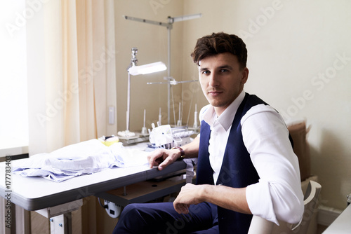 Plakat Portret przystojna krawiecka patrzeje kamera podczas gdy pracujący z serger maszyną w nowożytnym atelier studiu