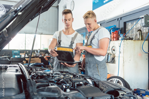 Zdjęcie XXL Doświadczony mechanik samochodowy używający laptopa do skanowania i interpretowania kodów błędów silnika obok praktykanta w nowoczesnym warsztacie samochodowym