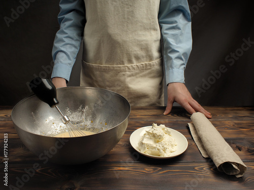 Plakat Przepis gotowania tiramisu, część trzecia: "Mieszanie masy jajka z mascarpone". Produkty na drewnianym stole: serek mascarpone, wstrząsana mieszanka żółtek jajecznych, trzepaczka do drutu.