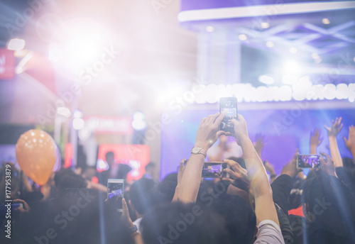 Plakat Azjatyckie kobiety trzyma mądrze telefon i stuka ekran dla brać obrazki na tłumie przy koncert sceną zaświecają, rozmyty tło