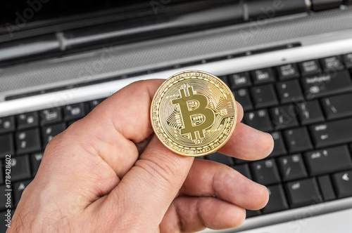 Man's hand holding metal coin of bitcoin over laptop keyboard.