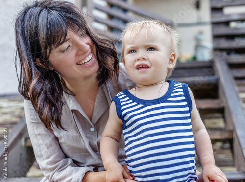 funny little boy one year with his mother, outdoor portrait. On the lawn next to the house, happiness, fun, family, rest