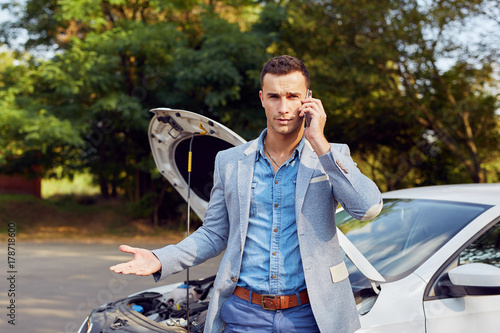 Young man stands next to a broken car calling