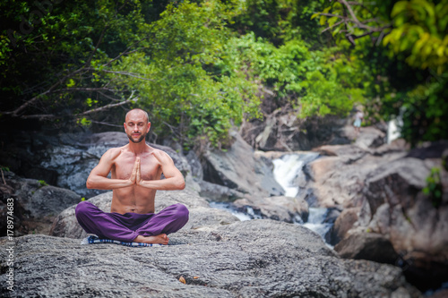 An attractive young man sits in a lotus pose and meditates against a background of stones and a waterfall.