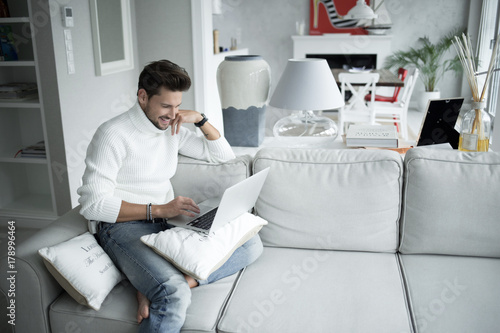 Plakat Przystojny mężczyzna ono uśmiecha się jego laptopu obsiadanie na leżance w domu